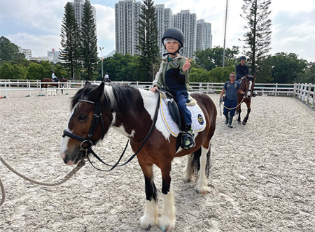 About 20 children from grassroots families visited the Tuen Mun Public Riding School in the company of civil service volunteers to sample the fun of horse riding. The Secretary for the Civil Service, Mrs Ingrid Yeung (second left), and volunteers from WSD helped a child get ready before the ride and another child enjoyed horse riding during the event.